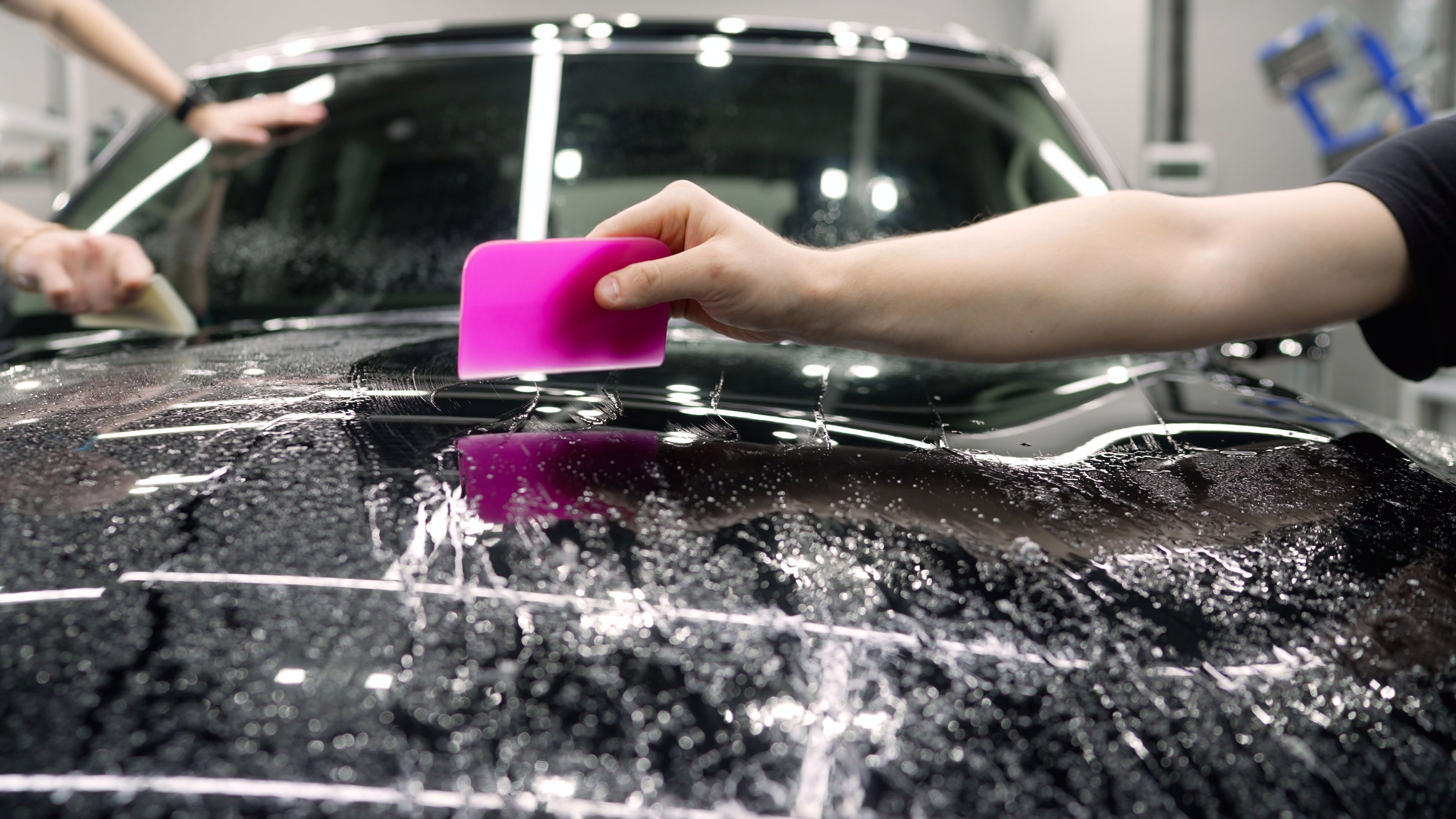 Smoothing the protective film on the car. The worker-detailer smoothes the protective film with the help of a working tool. Polymer protective outer coating, close-up. Applying a protective film.